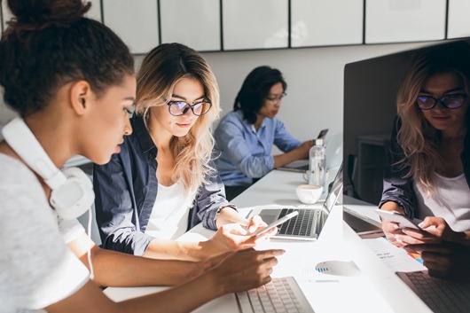 Pretty freelance copywriter texting message on phone while her colleagues typing on keyboards. Indoor portrait of young programmers of different ethnicities working in office..